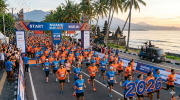 Thousands of marathon runners passing the start gate of Nuanu Run 2026 along a scenic Bali coastal road with a temple and mountains in the background.