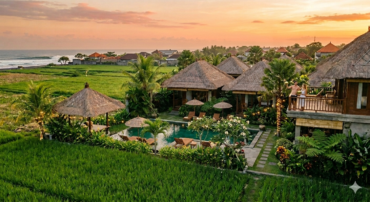 An aerial view of a traditional Balinese-style resort at sunset, featuring thatched-roof villas, a central swimming pool, and lush green rice paddies stretching to the ocean. A couple stands on a balcony.