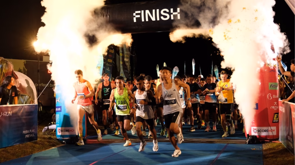 A group of marathon runners sprinting across the finish line at night with celebratory smoke machines and bright event lights.