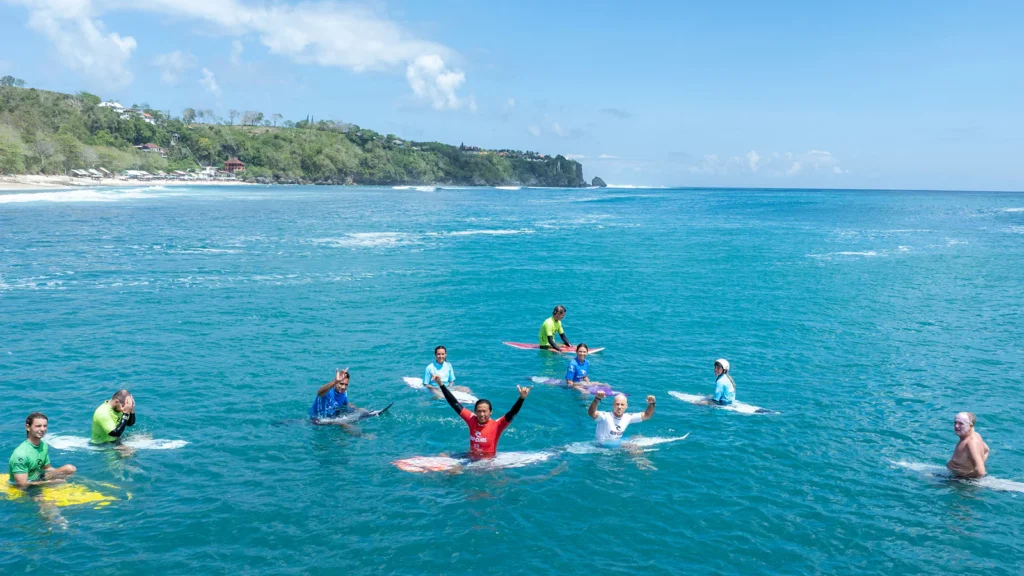 A group of professional surfers wearing event jerseys floating on their surfboards in the clear blue ocean during the Rip Curl Padang Padang competition in Bali.