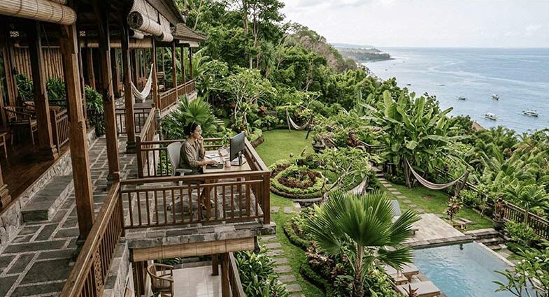 A female remote worker working on a desktop computer setup on a wooden villa balcony overlooking lush tropical gardens and the ocean.