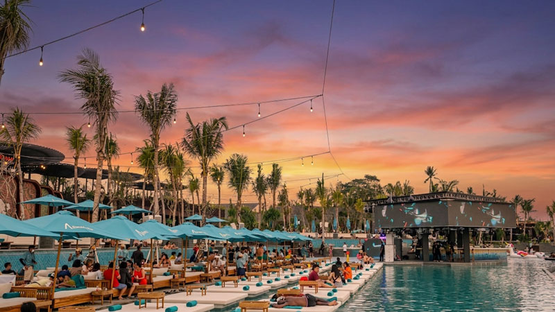 Visitors relaxing on daybeds lined up beside a large swimming pool at Atlas Beach Club in Canggu, Bali, under blue umbrellas and a vibrant orange sunset sky.