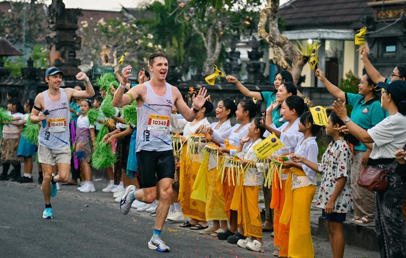 A male marathon runner smiling and waving as he passes local Balinese spectators and dancers dressed in traditional yellow attire cheering along the course.