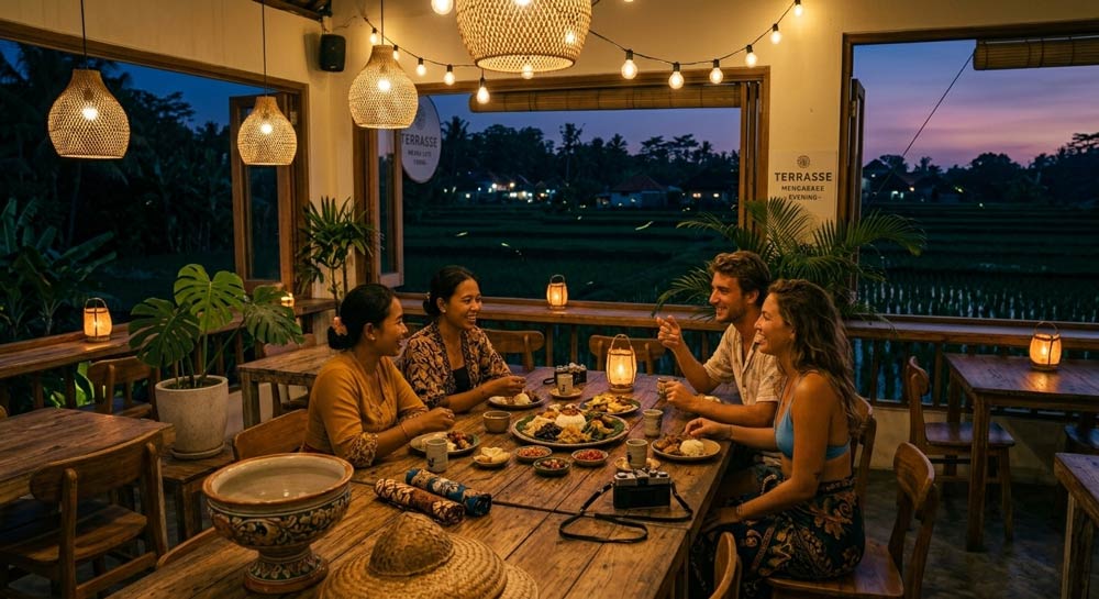 A group of diverse friends enjoying an authentic Balinese dinner together around a long wooden table in a warm, lantern-lit open-air restaurant overlooking rice fields at dusk.