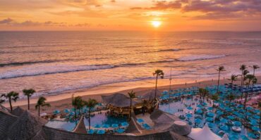 Aerial view of Finns Beach Club in Canggu, Bali, showing bamboo roofs, swimming pools with blue umbrellas, and crowds enjoying a sunset over the ocean shore.