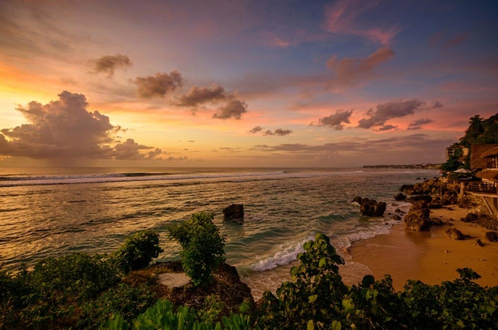 A breathtaking sunset over Bingin Beach in Bali, featuring golden skies, soft ocean waves, rock formations, and beachside villas along the cliffside.