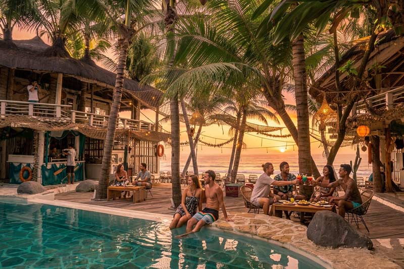 A group of friends enjoying drinks around a pool deck at La Brisa Beach Club in Canggu, Bali, surrounded by rustic wooden structures, palm trees, and a warm sunset over the sea.
