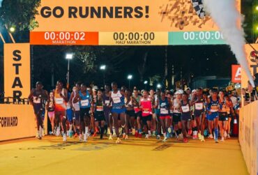 Elite long-distance runners crossing the yellow carpeted starting line of a marathon event under a large digital timer gate with smoke flares.