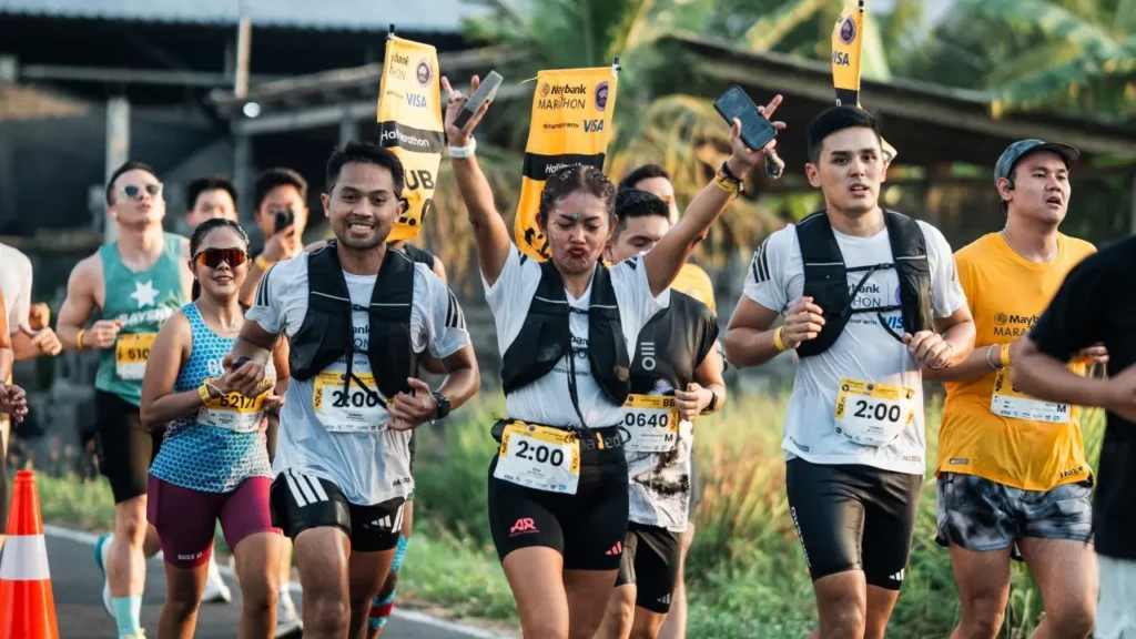 Official 2:00 target pacers with yellow flags attached to their backs running alongside participants during the Maybank Marathon event in Bali.