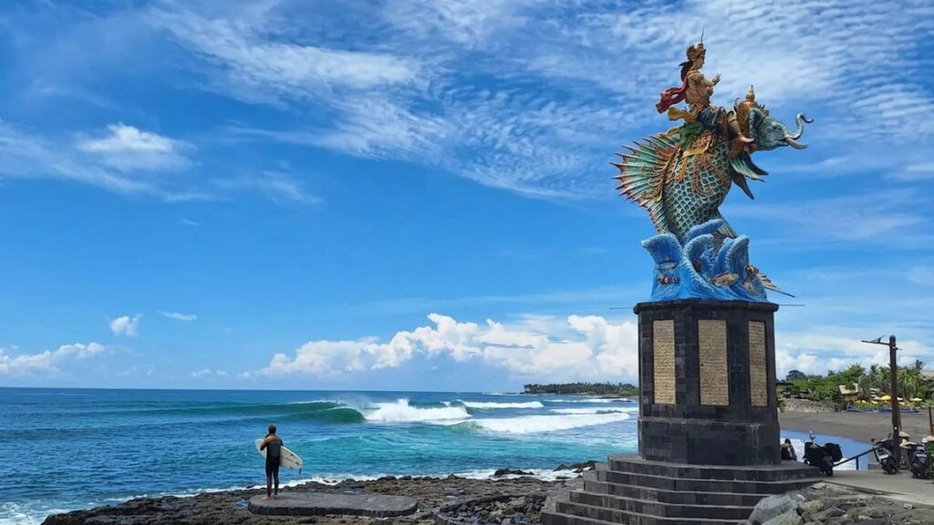 A surfer standing on a rocky shore holding a surfboard, looking at ocean waves next to a large Balinese Gajah Mina statue under a bright blue sky.