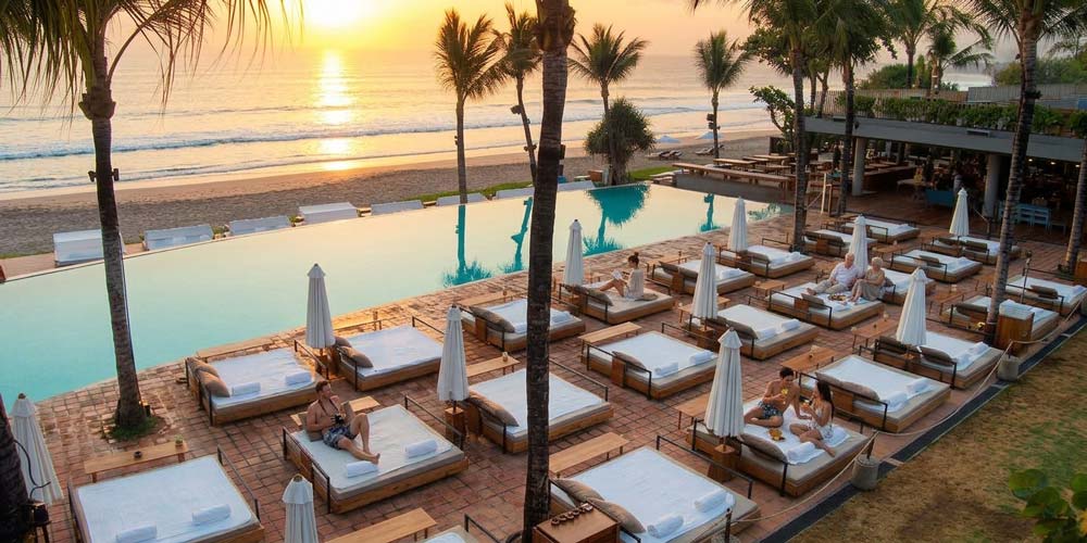 High-angle view of a luxury beachfront day club and pool resort in Bali, featuring rows of sunbeds, palm trees, an infinity pool, and guests relaxing during sunset.