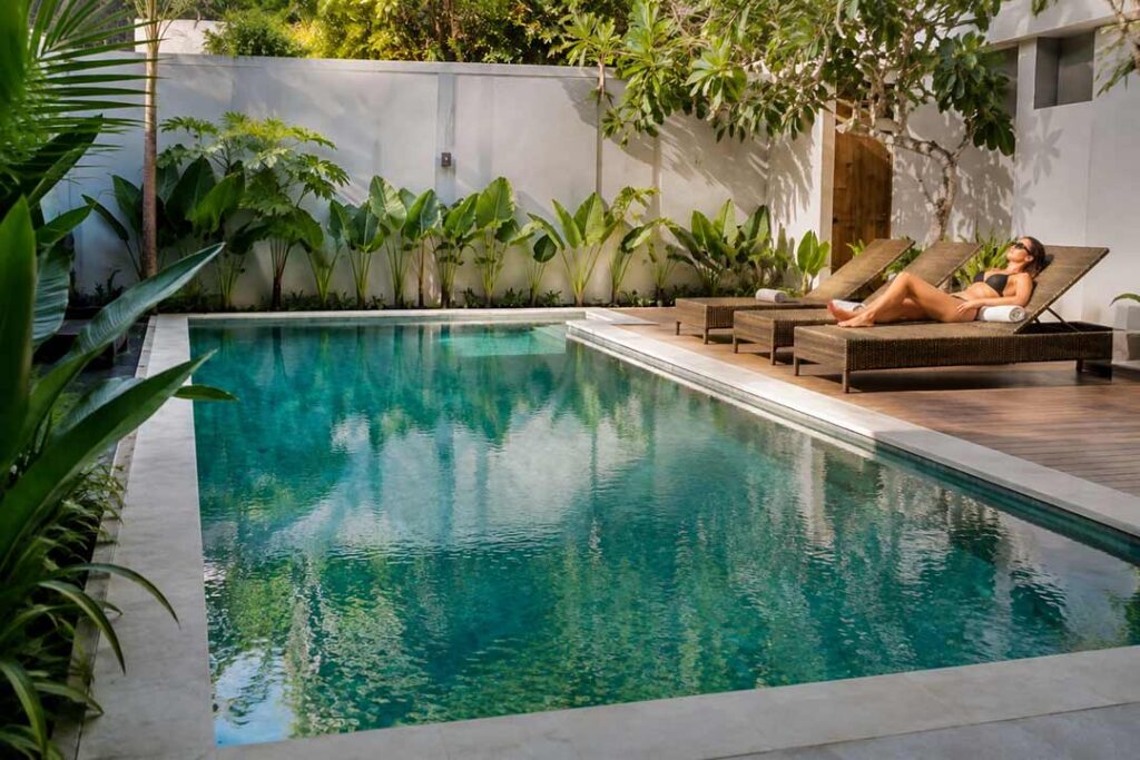 A woman in a black bikini sunbathing on a woven lounge chair beside a clear swimming pool surrounded by tropical green foliage.
