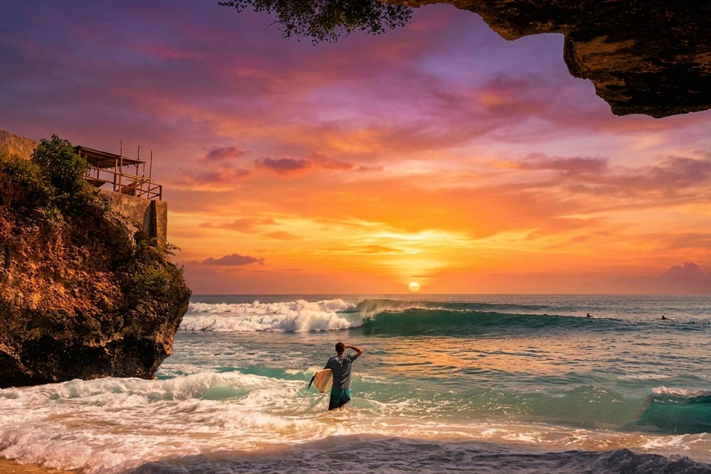 A male surfer holding a surfboard standing in shallow tide waves, looking out at a vibrant golden-orange sunset and rolling ocean waves framed by a cave cliff in Suluban Beach, Uluwatu, Bali.