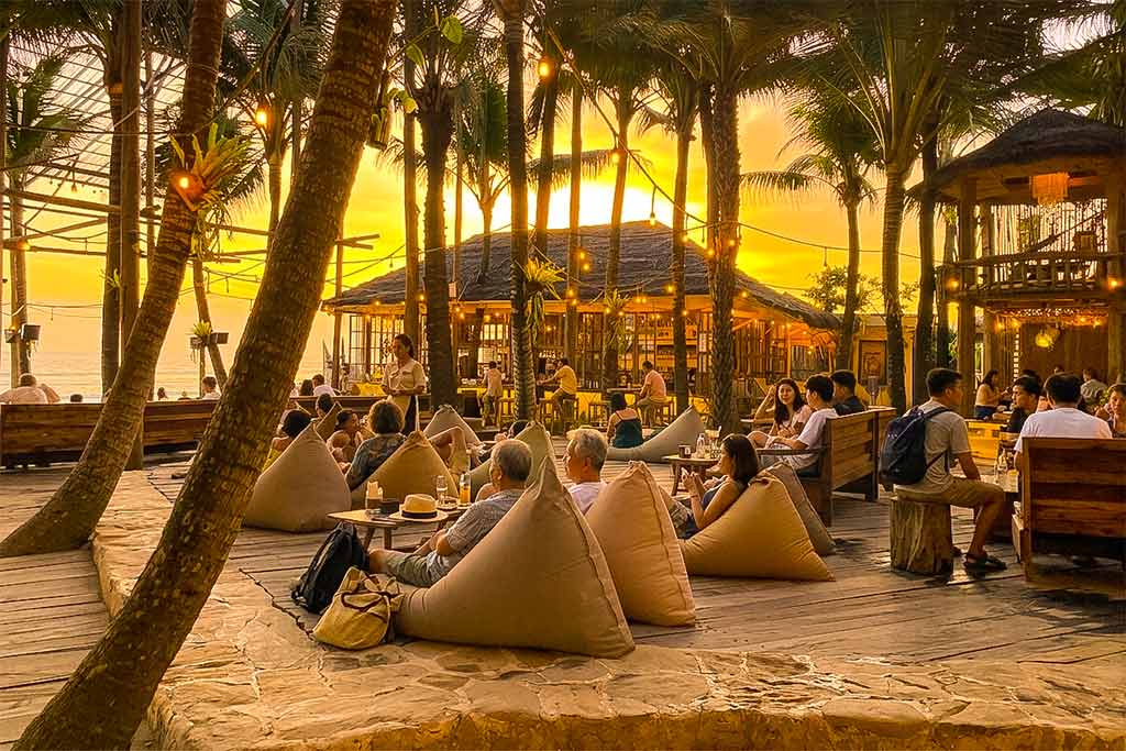 Asian travelers relaxing on cozy beanbags under palm trees at a beach cafe during their stay in Canggu.