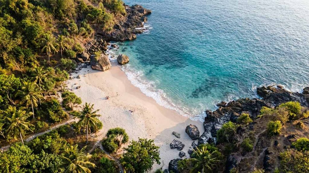 High aerial drone view of the secluded Bias Tugel Beach in Padangbai, Bali, showing a pale sandy cove bordered by dark volcanic rocks and lush tropical greenery at golden hour.