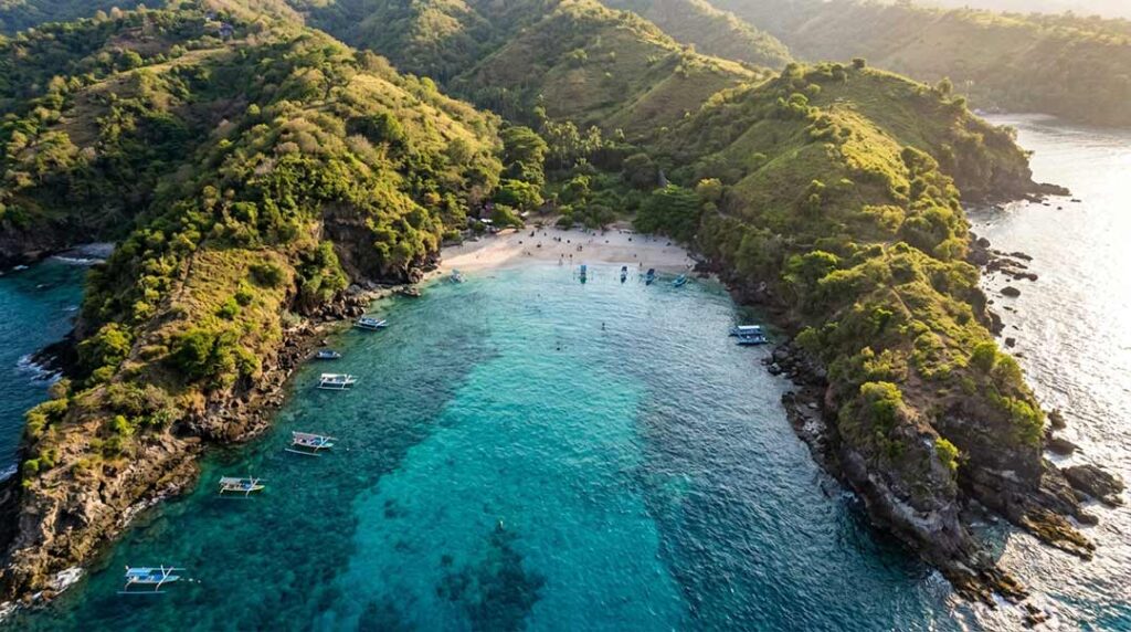 High aerial drone view of Blue Lagoon Beach in Padangbai, Bali, showing a sheltered cove with vibrant turquoise waters, surrounding green hills, and snorkeling boats during late afternoon.