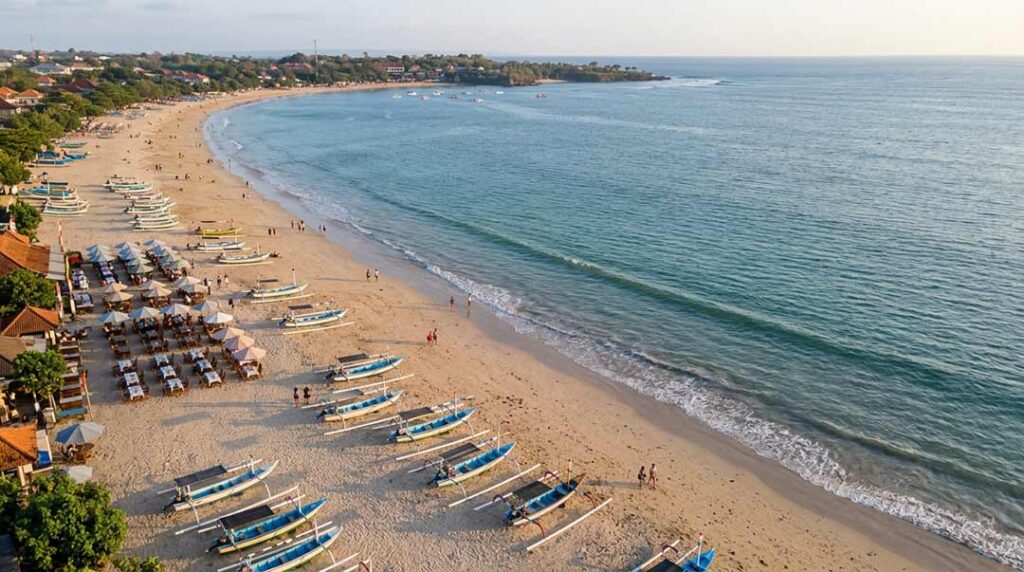 High aerial drone view of traditional jukung fishing boats lined up on the golden sand of Jimbaran Beach, Bali, near beachfront seafood restaurants during golden hour