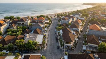 Low-altitude drone view of a paved street in Legian leading towards the sandy shore and ocean at Kuta Beach, Bali, during late afternoon.