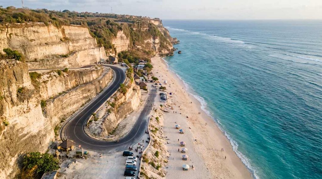 High aerial drone view of white limestone cliffs and a winding paved road leading down to Melasti Beach, Bali, at golden hour.