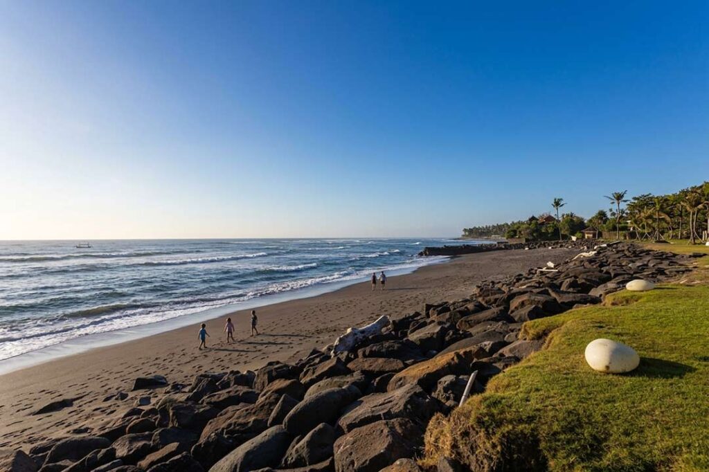 A serene black sand shoreline at Cemagi Beach, Bali, with distant families, a rocky barrier, and clear blue sky during sunrise.