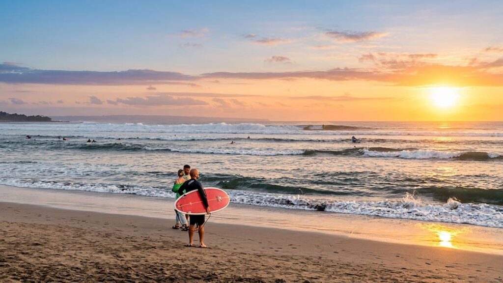 A man holding a red and white surfboard stands on a beach in Mengwi, Bali, with other surfers in the background and a sunset over the ocean.