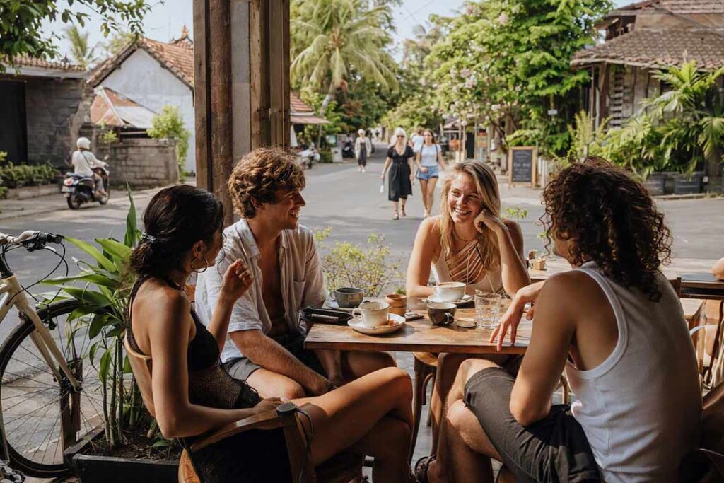 Four young travelers laughing together over coffee at an open-air tropical café in Canggu, Bali.