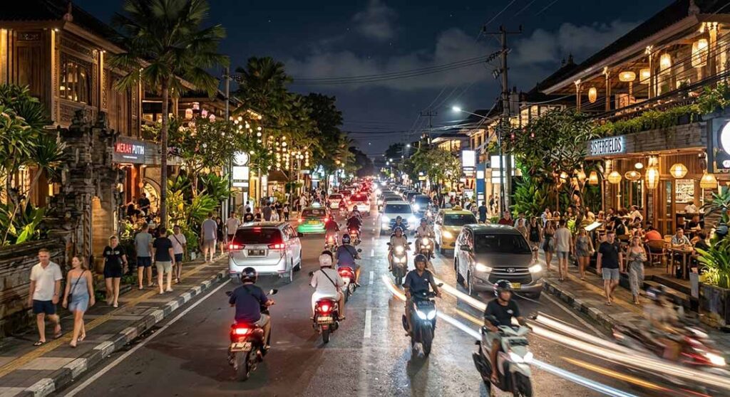 Busy night street scene in Seminyak, Bali, with scooters driving past brightly lit shops and restaurants.