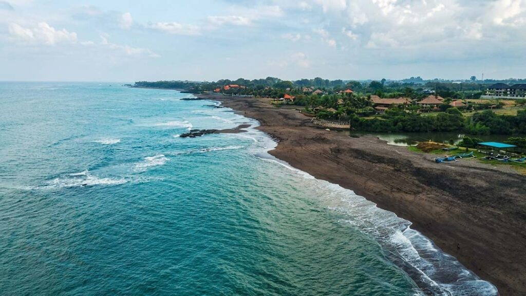 An aerial drone shot showing turquoise ocean waves crashing against the dark volcanic shoreline of Seseh Beach Bali surrounded by lush greenery and local coastal villas.