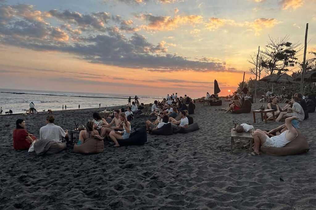 A crowd of people relaxed on beanbags and at tables on the dark volcanic sand of Seseh Beach, Bali, watching a spectacular sunset over the ocean.