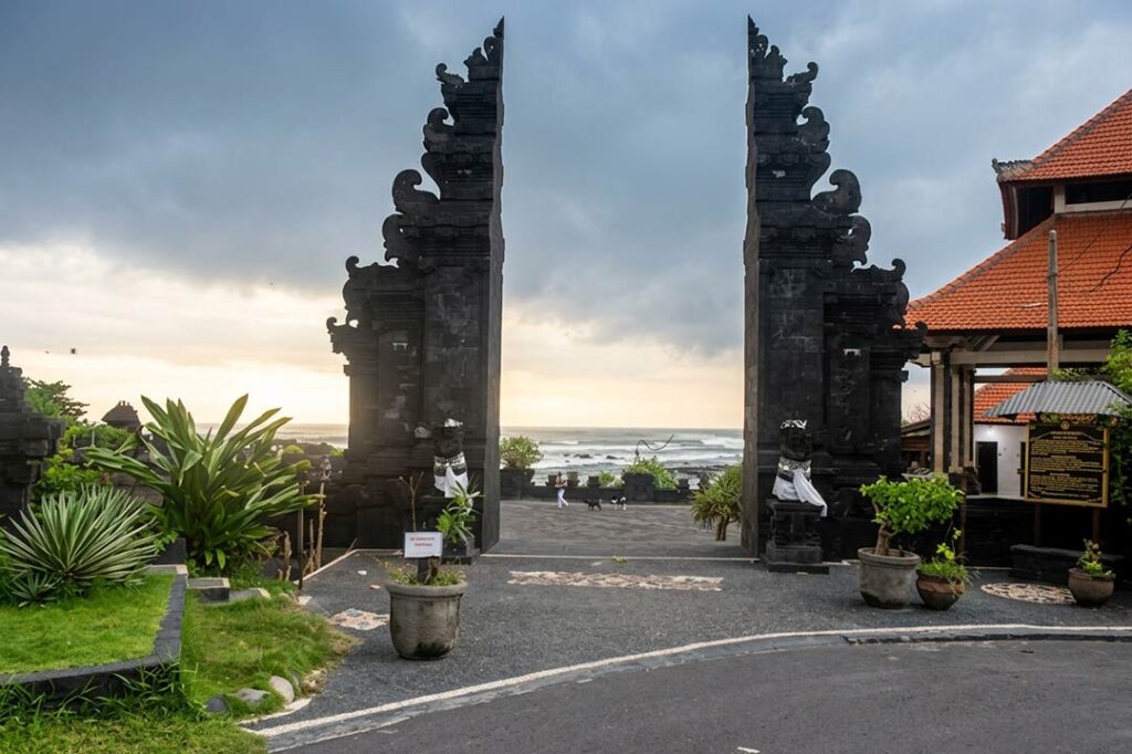 A traditional Balinese Hindu split gateway (Candi Bentar) at sunrise, with a clear view of the ocean and horizon at seseh beach bali, near a beachside temple complex.