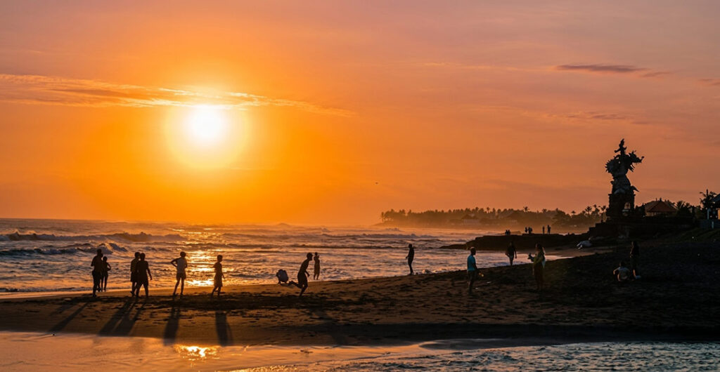Silhouetted people and a large dragon statue on a dark sand beach in Bali during a golden sunset.