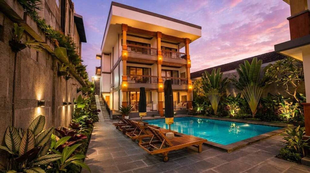 Elegant outdoor pool view at The Tunjung Boutique Resort with wooden sun loungers, closed black umbrellas, lush tropical plants, and a four-story room building under a twilight sky.
