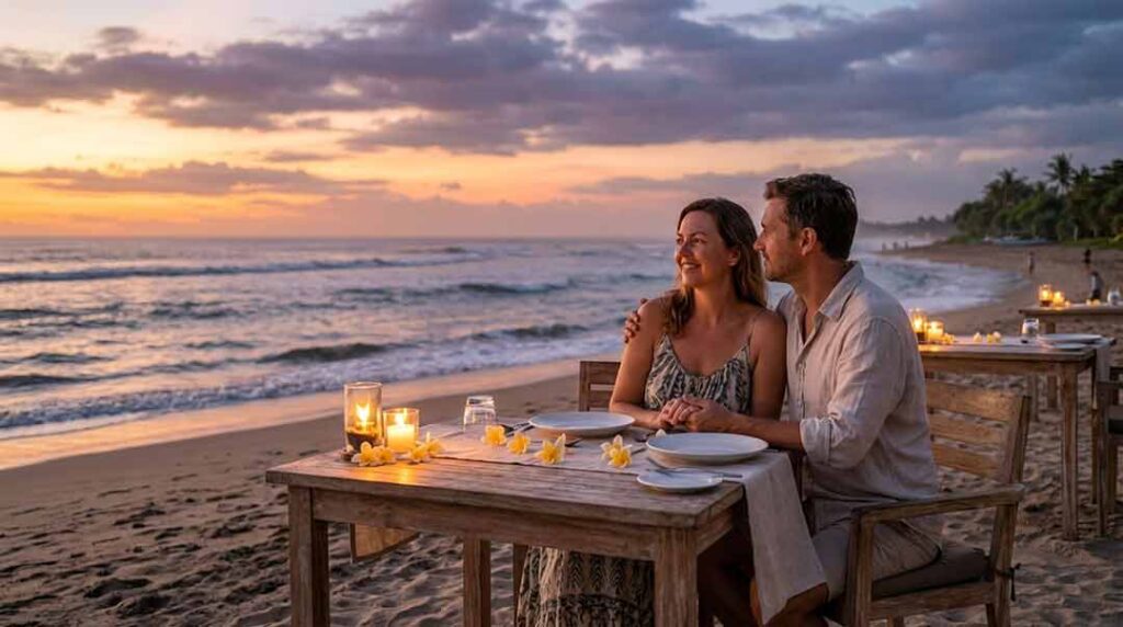 A couple sitting together at a candlelit wooden table on a sandy Bali beach enjoying dinner during sunset.