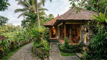 A quiet inland Balinese boutique villa with traditional thatched roofing, natural stone paving, and lush tropical greenery along a cobblestone lane.