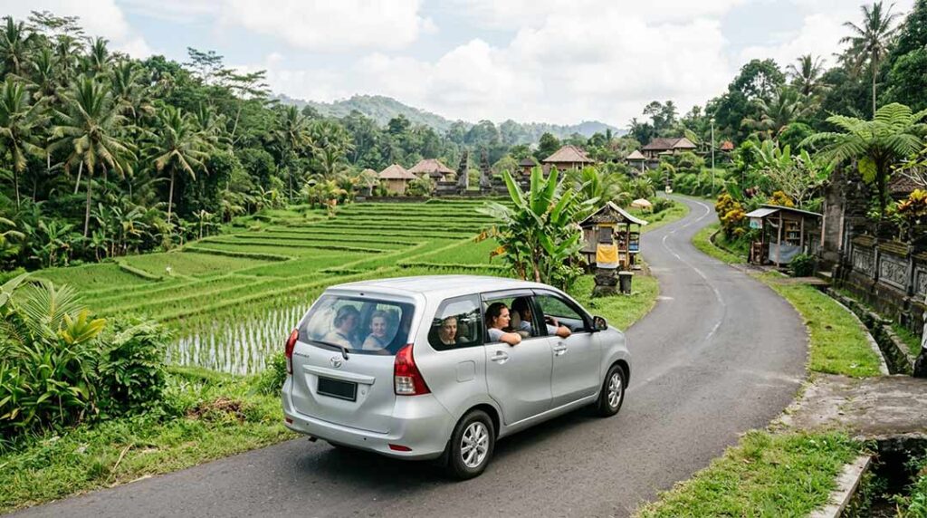 A silver passenger car carrying travelers along a winding inland road surrounded by green rice terraces and tropical trees in Bali.