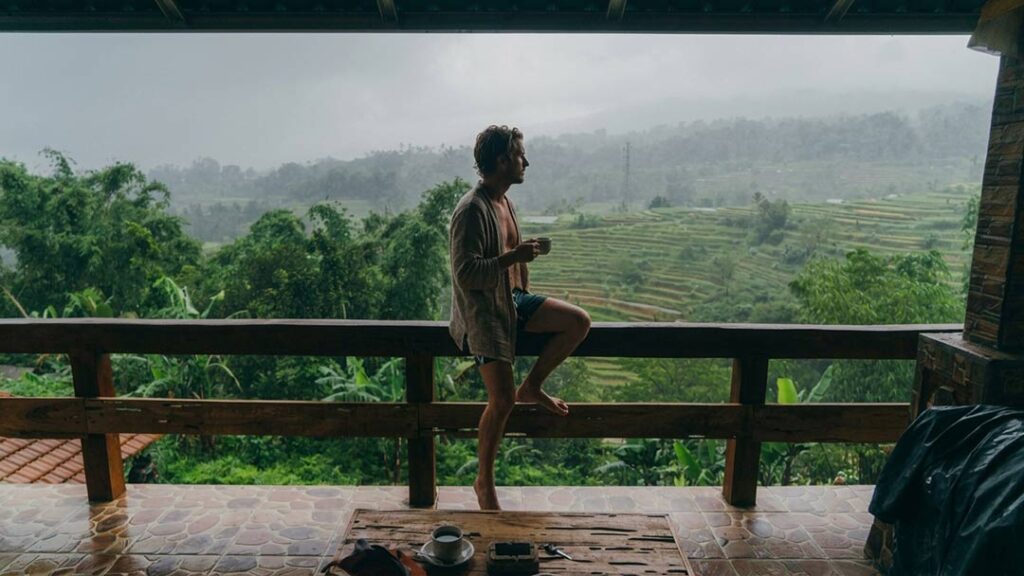 A man sitting on a wooden balcony railing holding a coffee mug while looking out at misty green rice terraces in Bali.