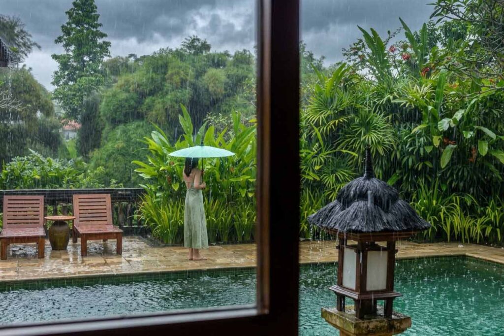 Woman with a light green umbrella standing by a swimming pool at a tropical villa in Bali during a heavy rain shower.