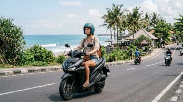 Tourist wearing a teal helmet riding a black scooter along a scenic coastal road in Bali with the ocean in the background.